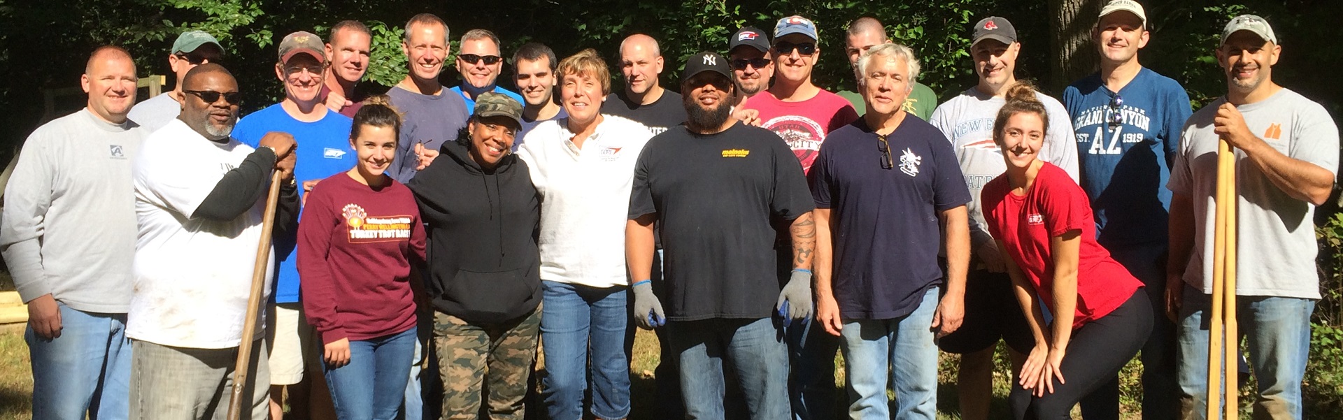 Many people gathered for the photo wearing white, red, black, and blue colored T-shirts with blue jeans, with 2 of them holding shovels.